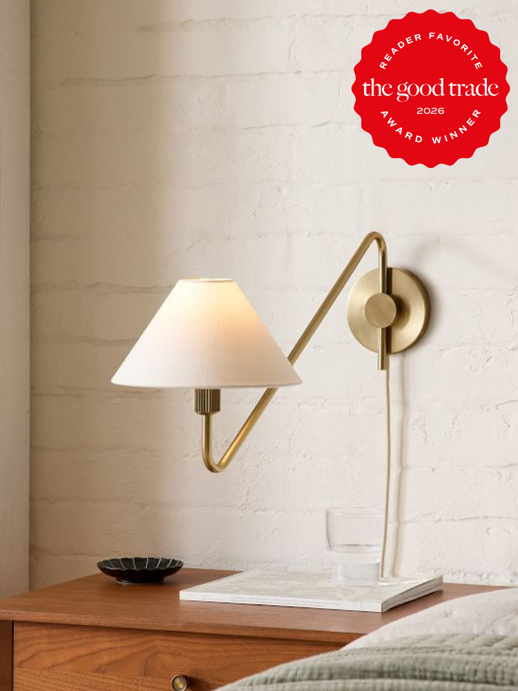 A modern brass wall lamp with a white shade is mounted above a bedside table holding a glass of water and a dish. A red award badge from The Good Trade is in the top right corner.