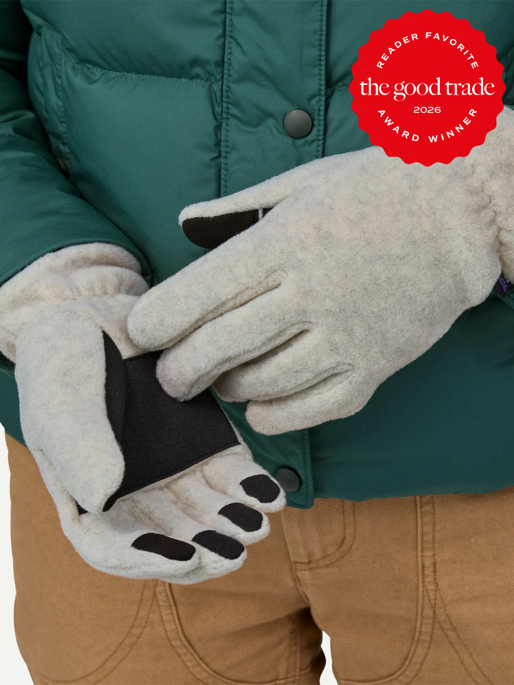 Person wearing light gray gloves with touch-screen compatible finger tips, holding one glove open; award badge in top right corner reads "The Good Trade Award Winner 2026.