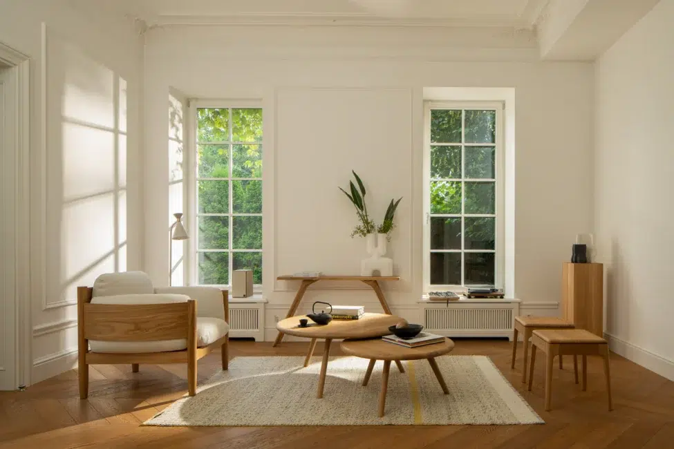 Minimalist living room with light wood furniture, a white armchair, nested coffee tables, a rug, large windows, and decorative plants, illuminated by natural sunlight.