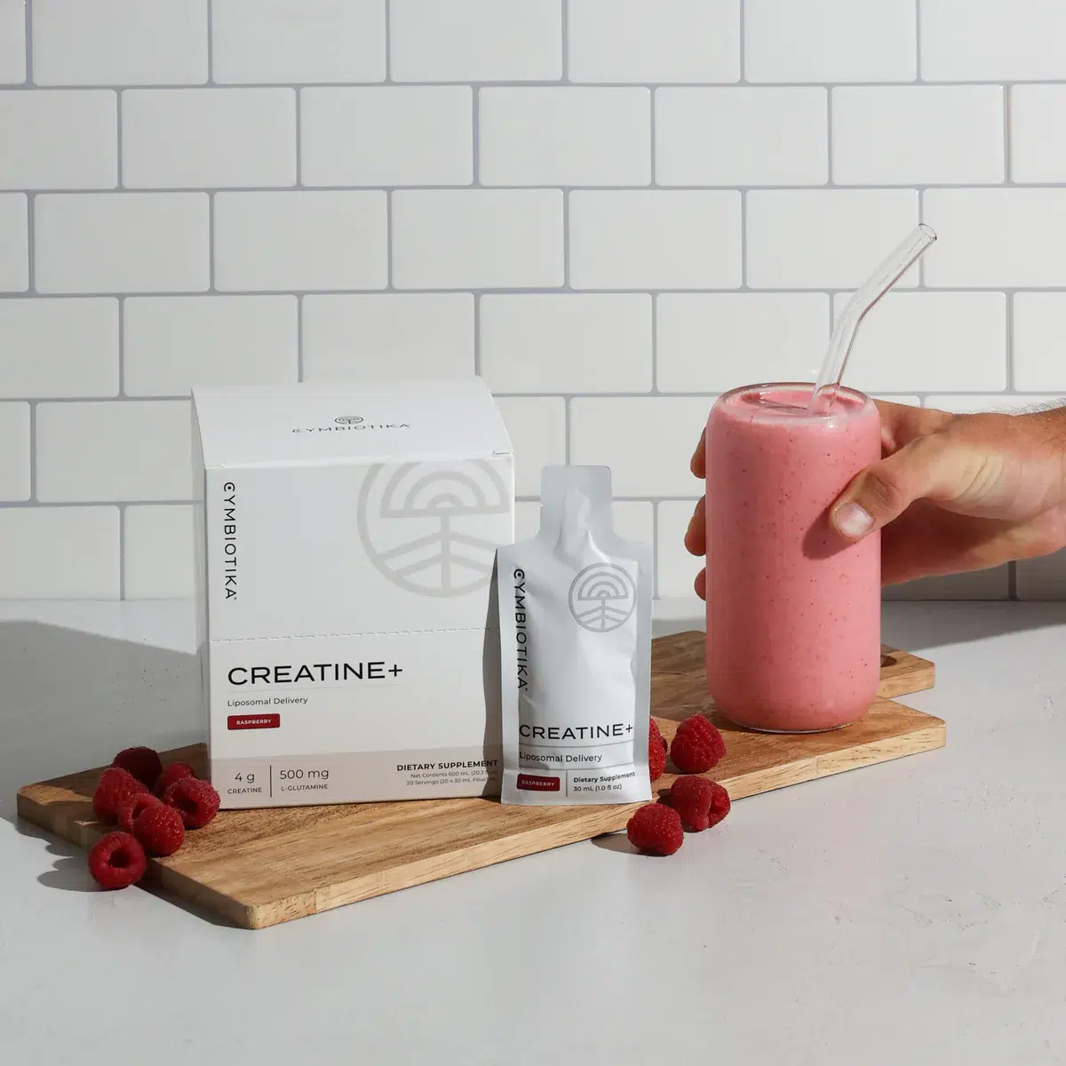Box and packet of creatine supplement next to a pink smoothie in a glass with a straw, surrounded by raspberries on a wooden board, against a white tiled background.