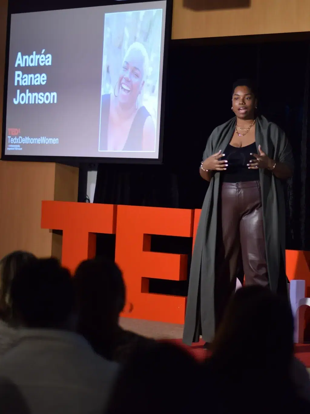 A speaker stands on a TEDx stage in front of two screens displaying the name “Andréa Ranae Johnson,” speaking to an audience.
