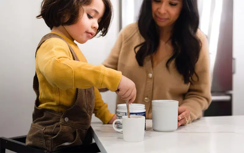 A child stirs a spoon in a white mug at a kitchen counter while an adult stands nearby with another mug.