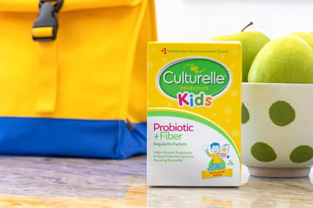 A box of Culturelle Kids Probiotic + Fiber stands on a counter next to a bowl of green apples and a blue and yellow lunch bag.