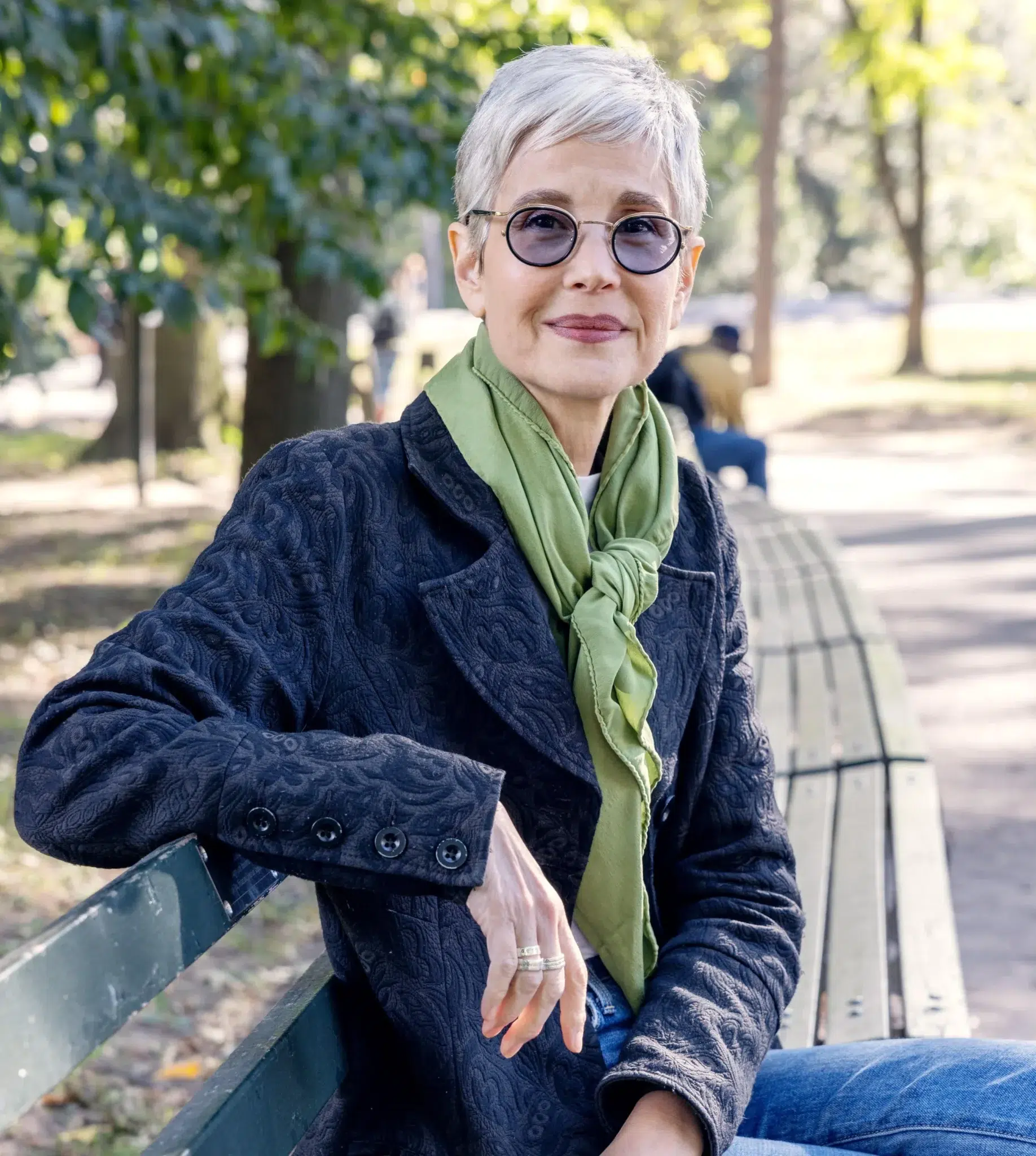 An older woman with short gray hair, glasses, and a green scarf sits on a park bench outdoors, looking at the camera.