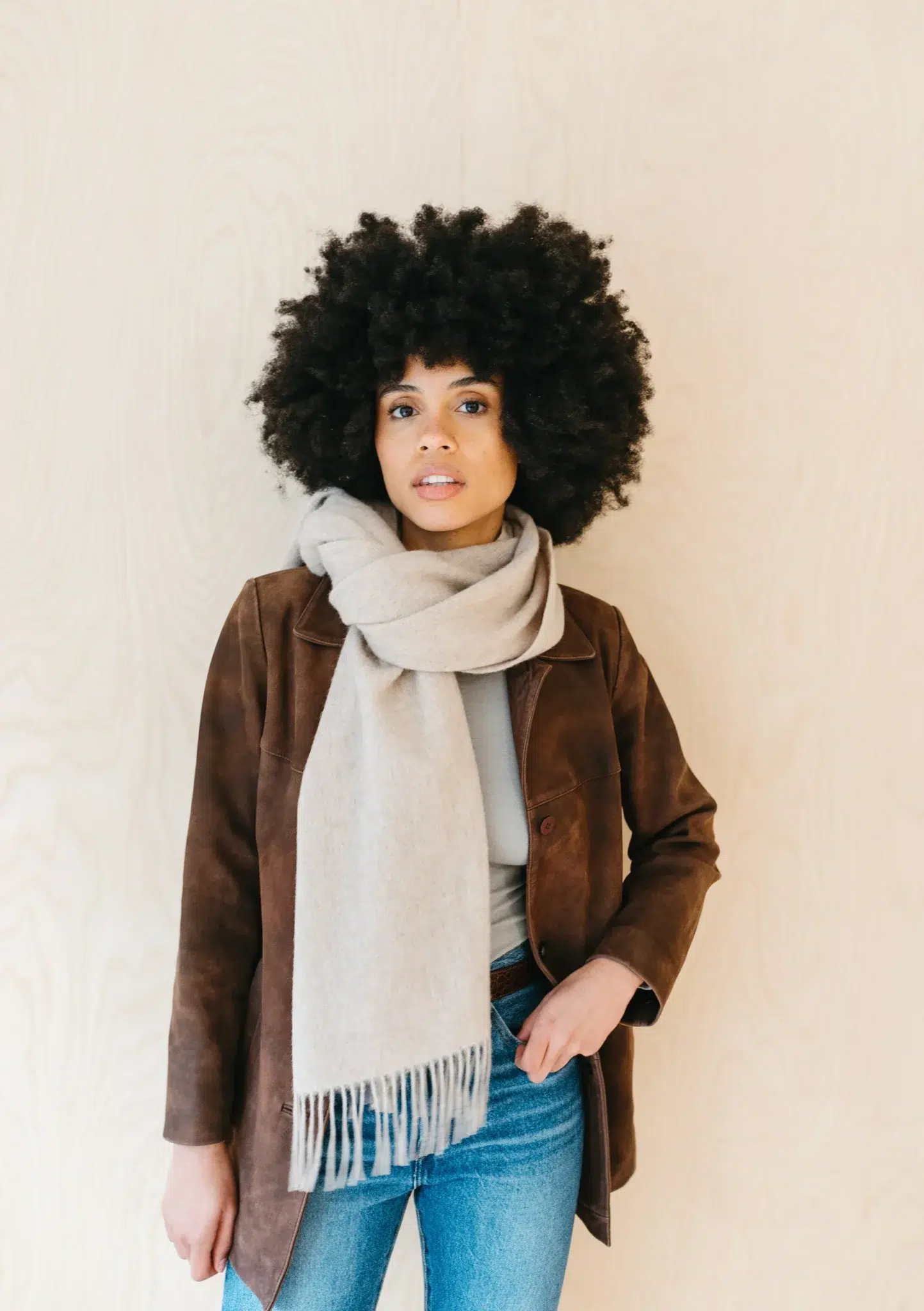 A woman with curly hair wears a brown jacket, beige scarf, and blue jeans, standing against a light-colored background.